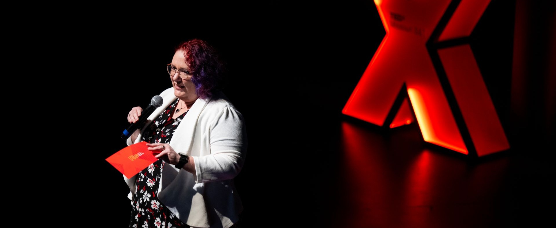 Lisa, a woman with two-tone red and purple dyed hair and wearing a white blazer, stands in front of a large red X. The spotlight is on her as she hosts the event.